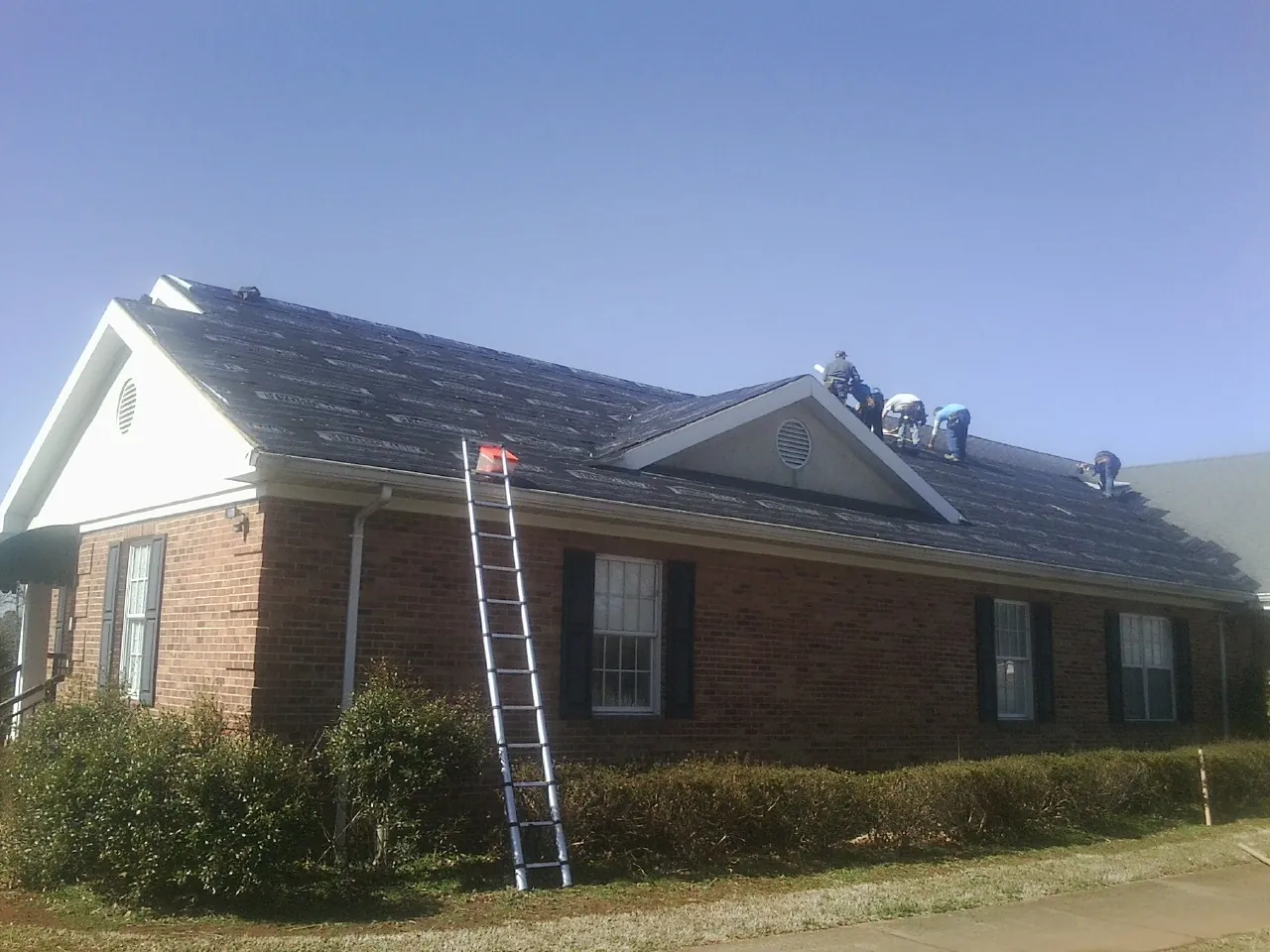 Crew tearing shingles off a brick ranch roof with a ladder set — Emergency Roof Repair work by our crew in Ninnekah