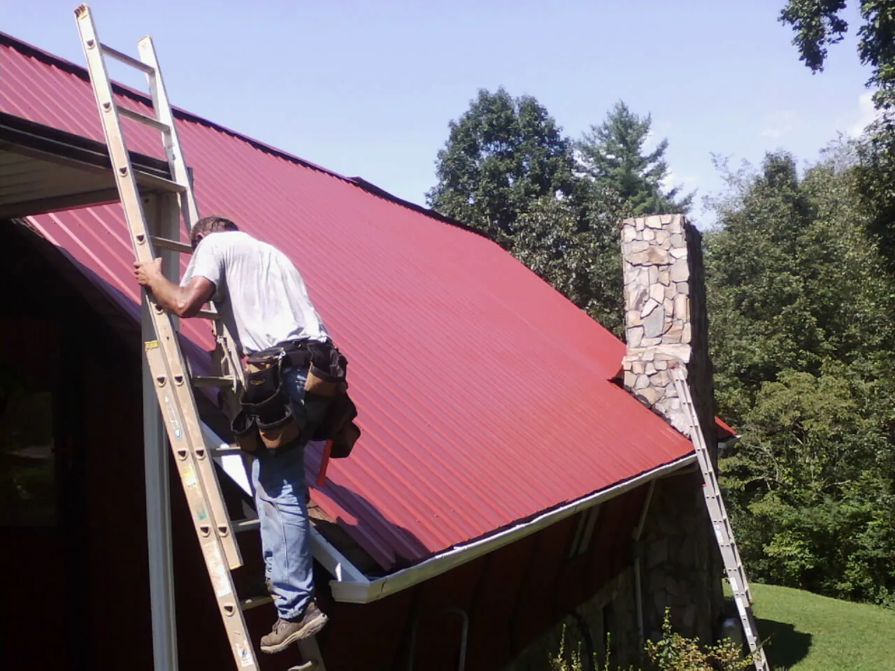 Roofer on a ladder beside a red metal roof and stone chimney — Soffit & Fascia Repair by Roofing Brothers in Ninnekah